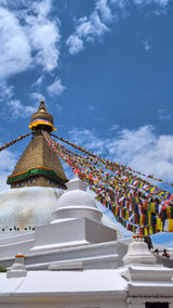 Colorful Tibetan prayer flags extend outward from the golden spire of the Boudhanath Stupa in Kathmandu, Nepal, under a bright blue sky with scattered clouds. The iconic Buddha eyes are visible on the stupa’s dome, surrounded by whitewashed architecture and visitors below.