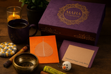 A warm lifestyle flat lay featuring a purple MAHA Treasures gift box surrounded by Nepali handmade items, including a brass singing bowl with an elephant design, an orange Lokta journal with a Bodhi leaf, rolled prayer flags, incense sticks, a felt snowman keychain, and a felt ball coaster, all arranged on a wooden surface with a lit candle and a cup of tea.