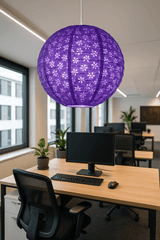 A vibrant purple Lokta paper lantern with floral cutouts hangs above a modern office desk with a computer monitor, keyboard, and black mesh chair, surrounded by large windows and indoor plants in a bright, minimalist workspace.