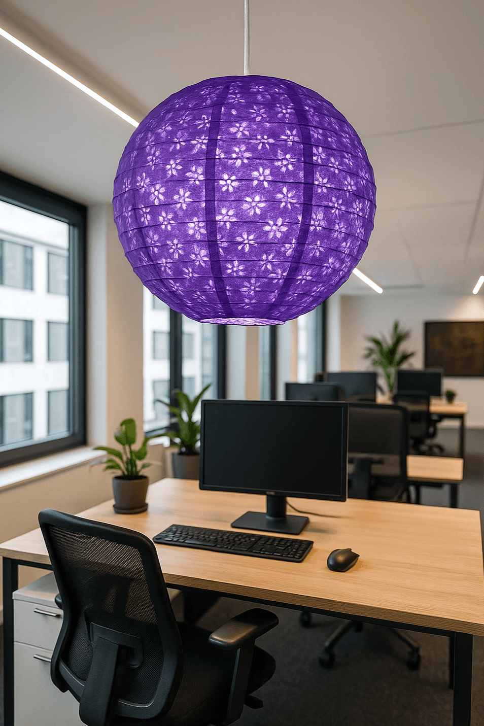 A vibrant purple Lokta paper lantern with floral cutouts hangs above a modern office desk with a computer monitor, keyboard, and black mesh chair, surrounded by large windows and indoor plants in a bright, minimalist workspace.