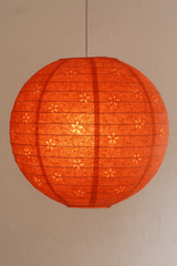 A handcrafted orange Lokta paper lantern with a floral cutout design hangs from a white cord, glowing warmly against a textured beige wall.