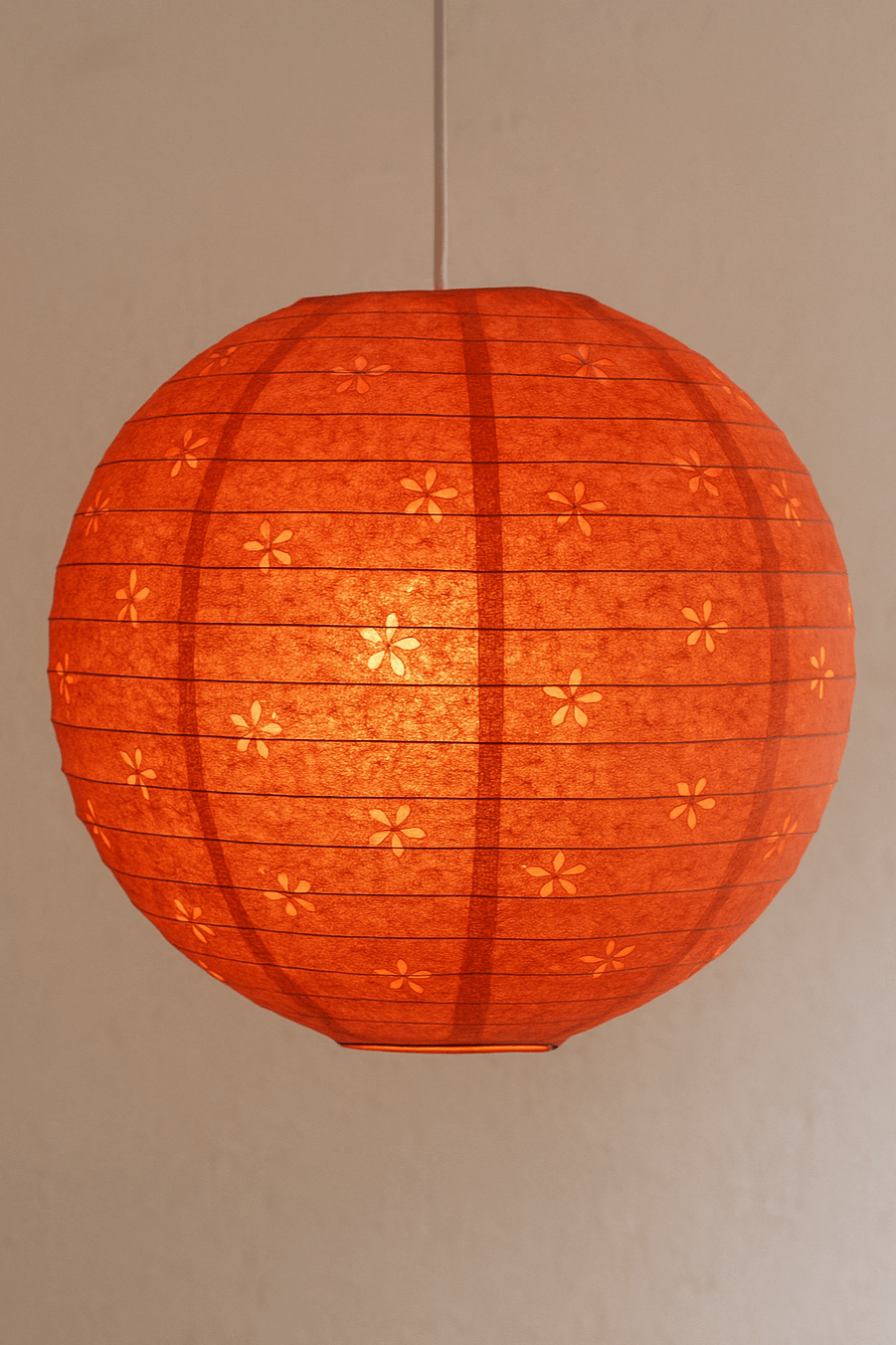 A handcrafted orange Lokta paper lantern with a floral cutout design hangs from a white cord, glowing warmly against a textured beige wall.
