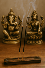 Two masala incense sticks burn in a wooden holder, releasing delicate trails of smoke, with brass statues of Ganesha and Lakshmi softly blurred in the background on a wooden altar.