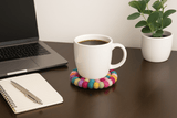 A photograph showcases a close-up of a neatly arranged workspace on a wooden desk, featuring a laptop, pen holder, and a colorful handmade felt ball coaster beside a cup.