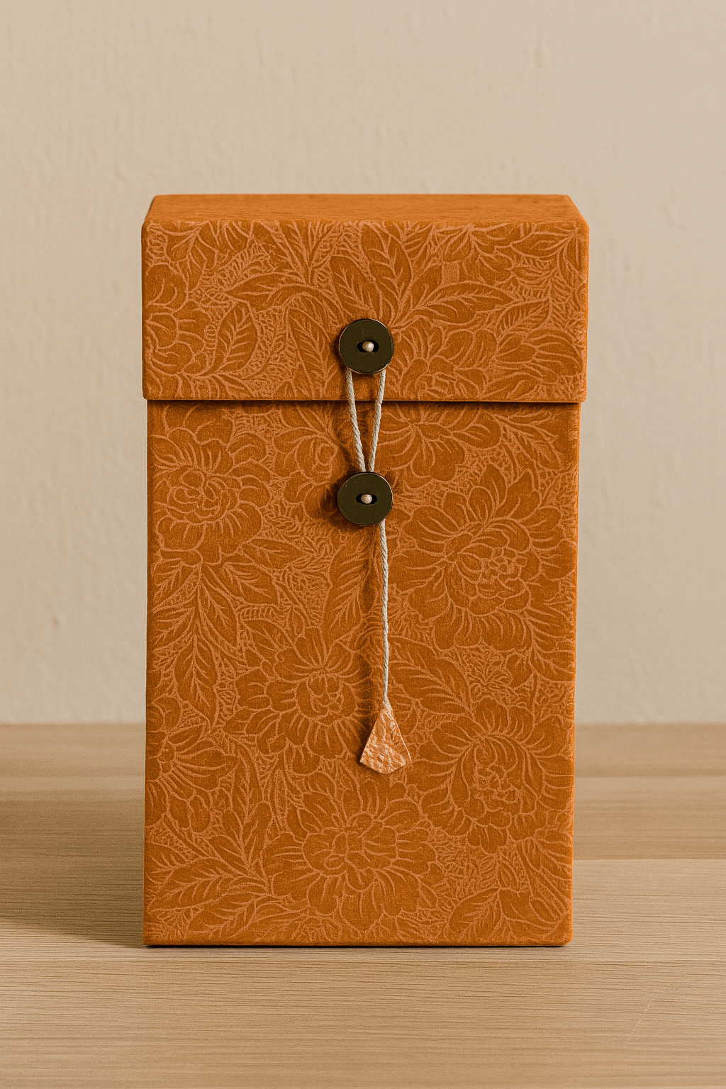 A rectangular Lokta paper box with floral patterns in a warm taupe hue, closed with a string wound around two round buttons, positioned against a simple beige surface.