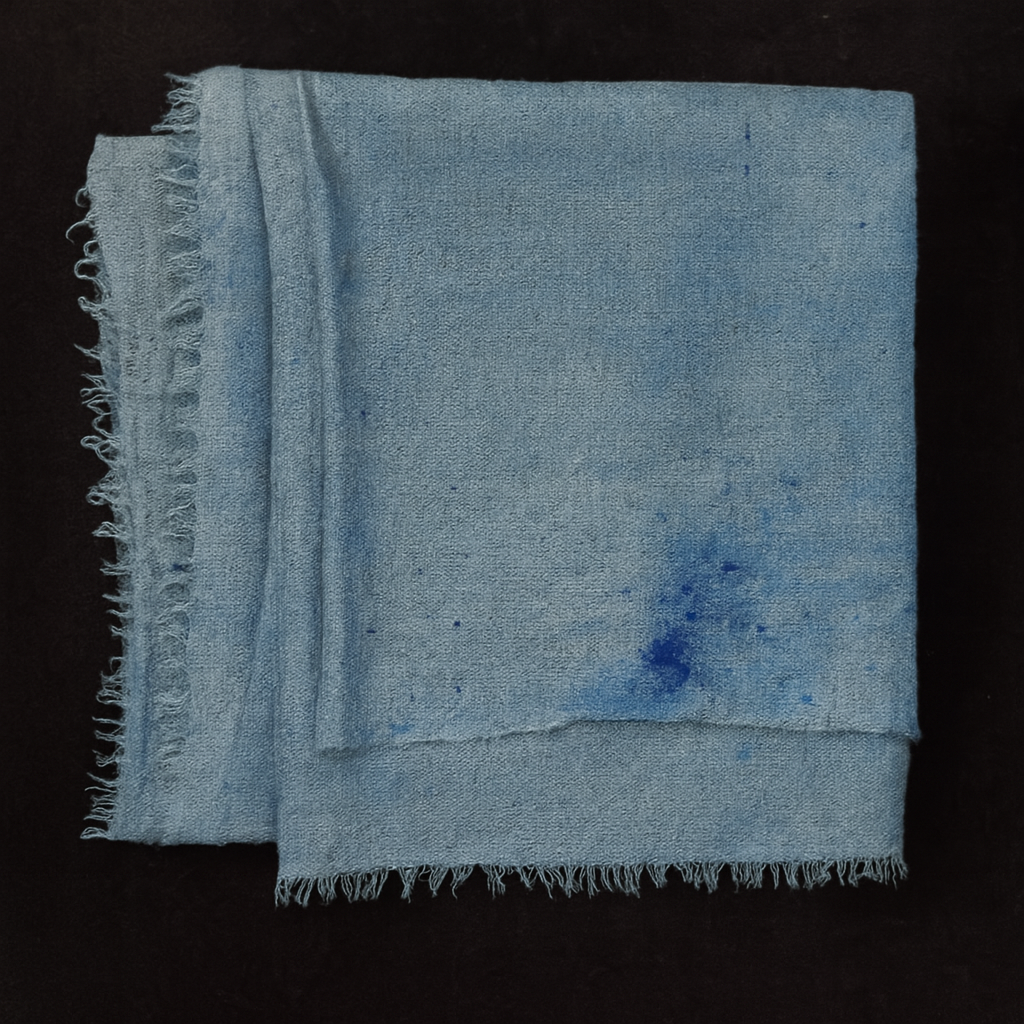 Folded handwoven pashmina stole in soft ice-dyed blue tones with subtle darker blue splashes, featuring delicate fringe edges, displayed on a dark fabric background.