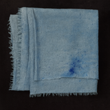 Folded handwoven pashmina stole in soft ice-dyed blue tones with subtle darker blue splashes, featuring delicate fringe edges, displayed on a dark fabric background.